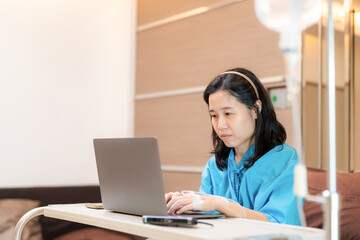 Asia patient woman sitting on the bed with saline solution in hospital ward using laptop, computer and check email. Working in clinic. Busy life of employee during sick. Healthcare and medical concept