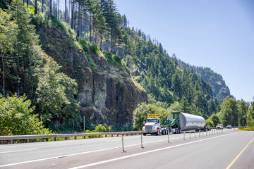Big rig semi truck with oversize load sign transporting long oversized windmill generator pole on specialized trolleys added to semi trailer running on the highway road in Columbia Gorge