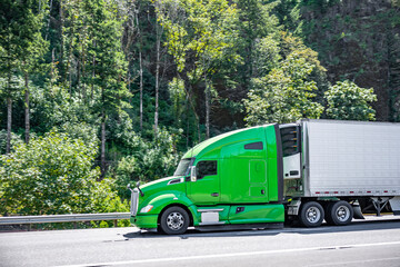 Fototapeta na wymiar Bright green industrial big rig semi truck transporting cargo in refrigerated semi trailer moving on the highway interstate road with forest on the side