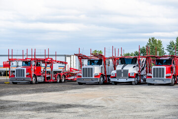 Bright red and white big rigs car haulers semi trucks with empty semi trailers standing on the industrial parking lot waiting for the next car loads