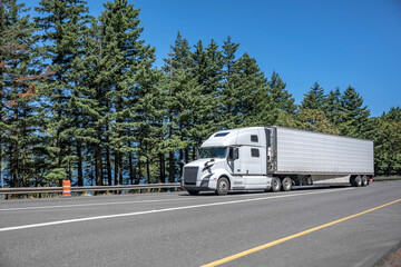 Comfortable pro white big rig semi truck transporting cargo in reefer semi trailer driving on the multiline highway road
