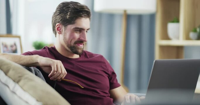 Young, casual and happy businessman on a laptop working remote at home in a modern living room. A smiling, relaxed and satisfied male feeling proud of his work or completed business project