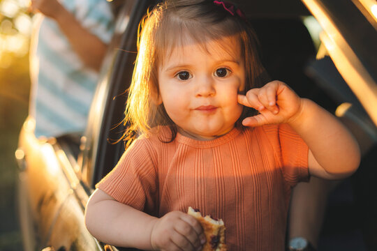 Baby Girl In The Car Looking Throw Window While Eating Tasty Pie. People Looking Outside. Childhood Concept.