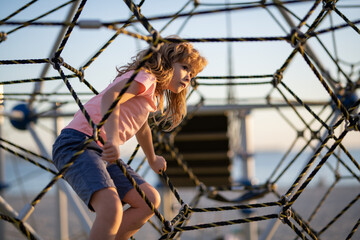 Kid boy climbing the net. Child playing on outdoor playground. Kids play on school or kindergarten yard. Kids play and climb outdoors on sunny summer day.