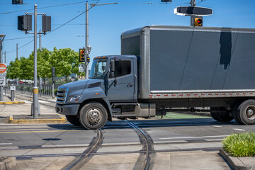 Middle duty day cab gray rig semi truck with box trailer transporting cargo running on the city street crossroad with traffic lights and tram rails.