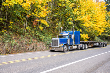 Fototapeta na wymiar Powerful classic big rig blue semi truck tractor with parts and empty flat bed semi trailer running on the narrow autumn road with yellow trees on the hill
