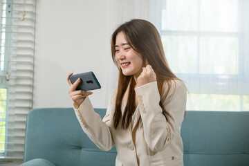 Fototapeta na wymiar Excited woman use mobile phone raising fist up she celebrating victory win online or received good news on sofa living room at home, Happy Asian female funny on smartphone feeling surprise excited