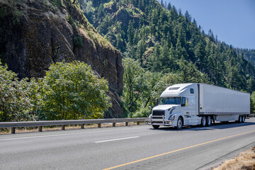 Powerful white big rig semi truck with grille guard transporting cargo in dry van semi trailer driving on the highway road with mountain and trees on the side