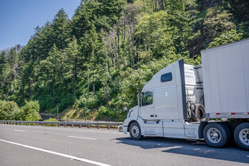 Professional big rig white semi truck with dry van semi trailer running on the international highway road with forest on the hillside