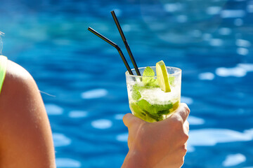 A close up of a glass of cool ice mahitto held by a tanned girl. Against the backdrop of a pool...