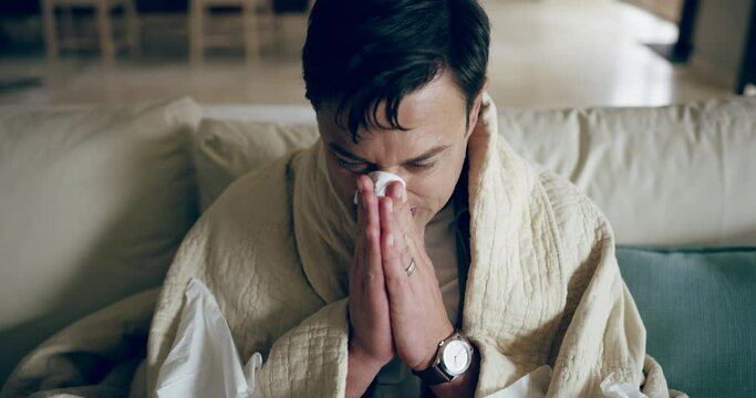 A sick or ill man blowing his nose suffering from the flu or cold sitting at home on a couch. Young unwell caucasian male with a fever or covid while quarantine in a house wrapped in a blanket