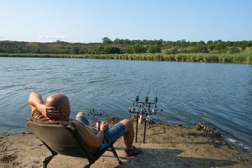Angler is fishing with carpfishing technique in a beautiful summer evening, using rod pod, bite alarms, swinger, rods. Fisherman with hot drink in mug waiting a bite with carp fishing equipment.