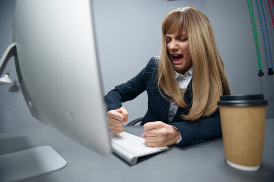 Evil irritated millennial blonde businesswoman worker in blue jacket breaks keyboard hate operation crash in gray modern office. Remote Job, Technology And Career Profession Concept. Wide angle