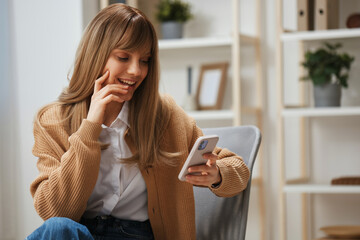 Cheerful young blonde woman in warm sweater get good news on studies by phone touch cheek sitting in armchair at home. Pause from work, take a break, social media in free time concept. Copy space