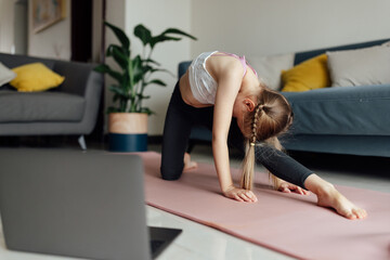 Naklejka premium Pretty caucasian little girl in sportswear watching online video on laptop and doing fitness exercises at home. Distant training with personal trainer, social distance or self-isolation, online