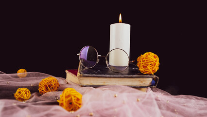 Burning candle books and thin veil with decor. Composition of burning wax candle near eyeglasses on stack of books near thin fabric with decorative wicker balls on black background