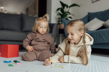 Little caucasian siblings drawing together on floor in living room at home. Happy girl eight years old and her baby sister painting with crayons. Art and crafts for toddler and preschooler
