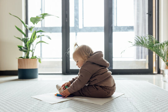 Cute caucasian baby drawing on floor in living room with potted green plant at home. Happy girl painting with crayons. 