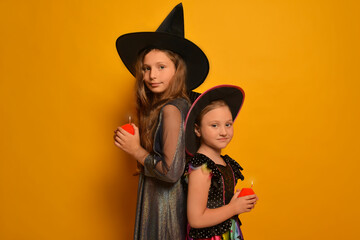 Cute little girls in witches costumes holding red ignited candles or burning candles in hands and looking at camera on a yellow background.

Two sisters on Halloween, kids in carnival costumes.