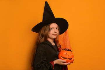 Cute little girl in wizard costume holding small pumpkin with spider and looking at camera on orange background. Cheerful witch child preparing for Halloween holiday in carnival costume.