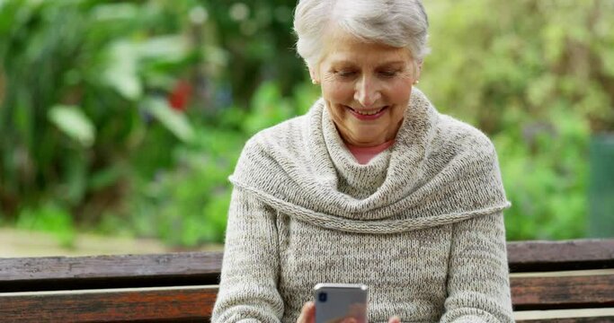 Cheerful and retired elderly female taking a selfie on park bench outside. A mature caucasian lady receiving good news text message from her loved ones. Smiling senior woman looking at phone.