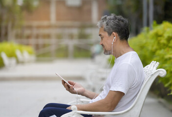 senior man with grey hair and beard in sportswear sitting on bench after workout feeling calm with headphones listening to music in the park, concept elderly people lifestyle, wellbeing