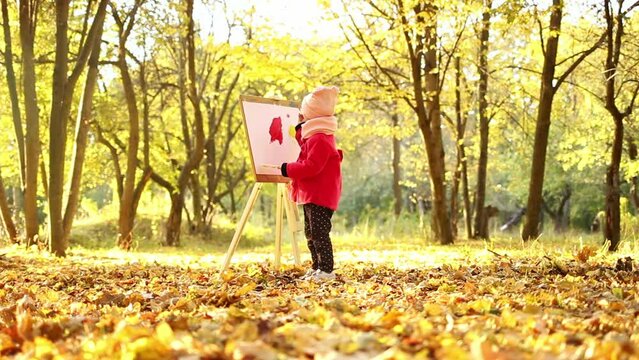 A little girl in a red coat paints an watercolor on easel in the park against the background of the autumn landscape. A cute child is taking paint from the palette, looks and smiles into the frame