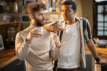Two guys of different ethnicity having warm conversation while drinking coffee on kitchen at home. Concept of close male friendship or relationship as gay couple. Caucasian and hispanic man together