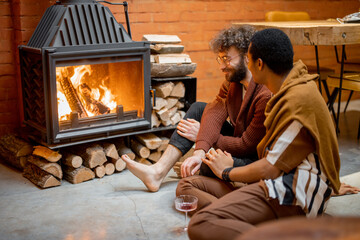 Two men talking while sitting together by the burning fireplace at cozy home. Concept of homosexual...