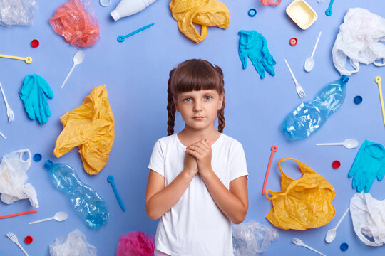 Indoor shot of cute sad little girl wearing white t shirt posing against blue wall and much plastic garbage around, keeps hands together, worries about our planet pollution.