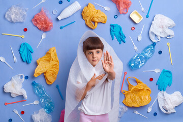 Image of confident little girl posing against blue wall and much plastic garbage around, showing...
