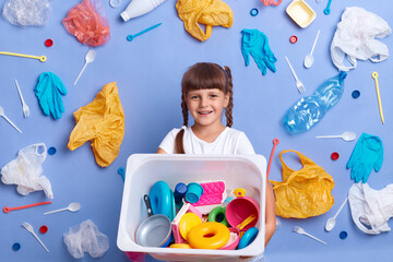 Smiling kind little girl wearing white t shirt posing against blue wall and much plastic garbage...
