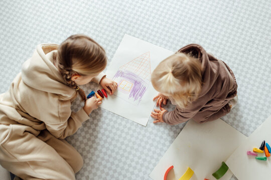 Little caucasian siblings drawing together on floor in living room at home. Happy girl eight years old and her baby sister painting with crayons. Art and crafts for toddler and preschooler