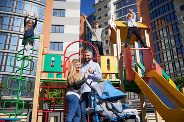 Three cheerful children playing on playground equipment with parents below. Front view of happy parents, holding thumb up, smiling with kids which are on top of slide outdoor. Concept of family.