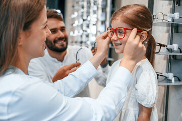 Family is together. Woman, man and little girl is choosing right glasses for correcting vision