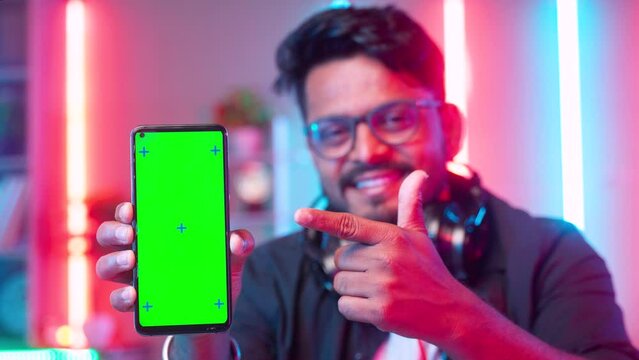 focus on smartphone, Professional gamer with headset showing green screen mobile phone by pointing finger while looking camera at home - concept of champion,app advertisement and champion.