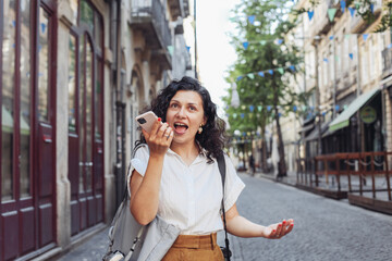 Young brunette woman emotionally recording a voice message with her smartphone, walking on an European street 