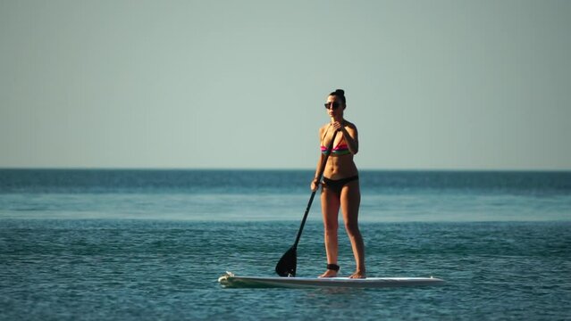 Sea woman sup. Silhouette of happy middle aged woman in rainbow bikini, surfing on SUP board, confident paddling through water surface. Idyllic sunset. Active lifestyle at sea or river. Slow motion