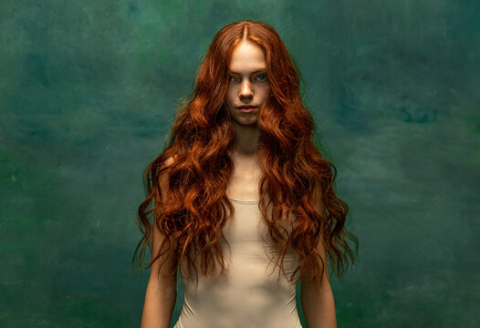 Adorable tender redhead girl with long curly hair isolated over dark green background. Fabolous curly hair and harming gaze. Concept of beauty, art