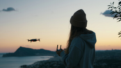 Soft view of young female, girl silhouette holding, using joystick to control quadcopter, flying drone outside on stunning sunset windy natural background to take pictures, videos for travel blog