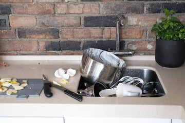 A pile of dirty dishes in a metal sink, in the kitchen, against a brick wall.