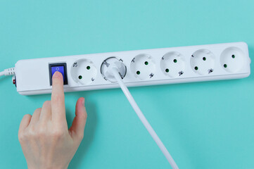 White surge protector with sockets.  hand presses the power button. Turquoise background.