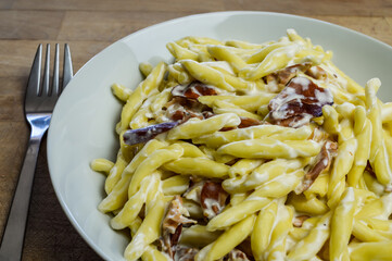 strozzapreti cream and speck, specialty from northern Italy, fresh twisted pasta with sauce made of fresh cream and speck, a smoked ham, and parmesan - in a white plate on a wooden cutting board