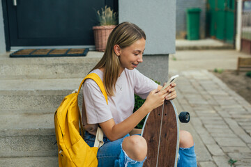 teenager with skateboard sitting on front step in front of her house and using mobile phone