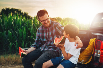 father and son eating watermelon sits on trunk of truck in field