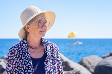 Attractive senior woman with hat and eyeglasses sitting at sea admiring beauty in nature, smiling...