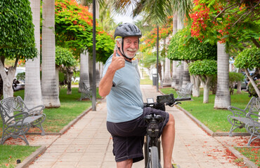 Happy active senior man cycling in the public park with electric bicycle looking back at camera with thumb up. Concept of healthy lifestyle and sustainable mobility