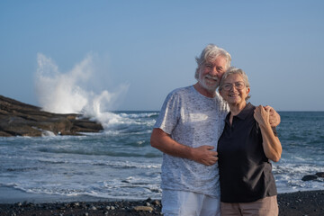 Beautiful happy caucasian senior couple standing on the beach at sunset looking at camera - smiling pensioners enjoying free time, sea vacation or retirement - Powered by Adobe