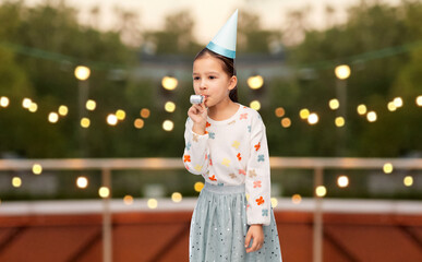 birthday, childhood and people concept - portrait of smiling little girl in party hat with blower having fun over garland lights on roof top background