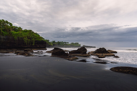 Black sand beach with ocean and waves in Balian, Bali. Beach with stones and cloudy sky